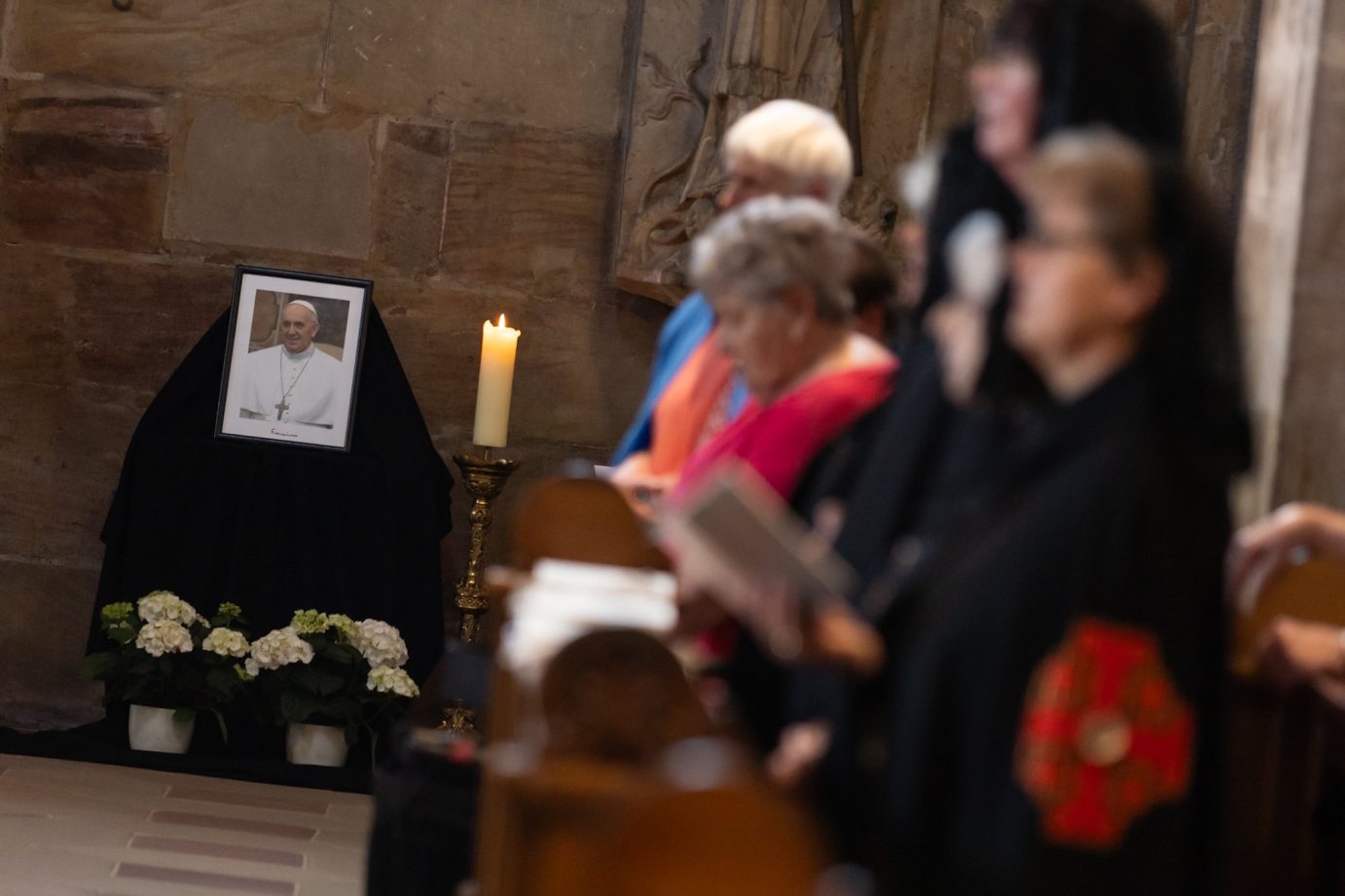 Das Bild zeigt im Hintergrund ein Bild von Papst Franziskus mit brennender Kerze und Blumen, im Vordergrund sind unscharf einige Gläubige zu erkennen.
