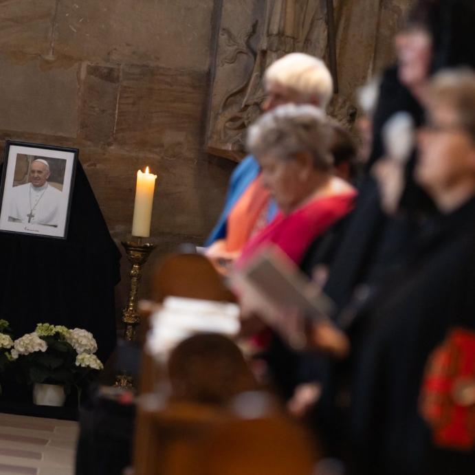 Das Bild zeigt im Hintergrund ein Bild von Papst Franziskus mit brennender Kerze und Blumen, im Vordergrund sind unscharf einige Gläubige zu erkennen.